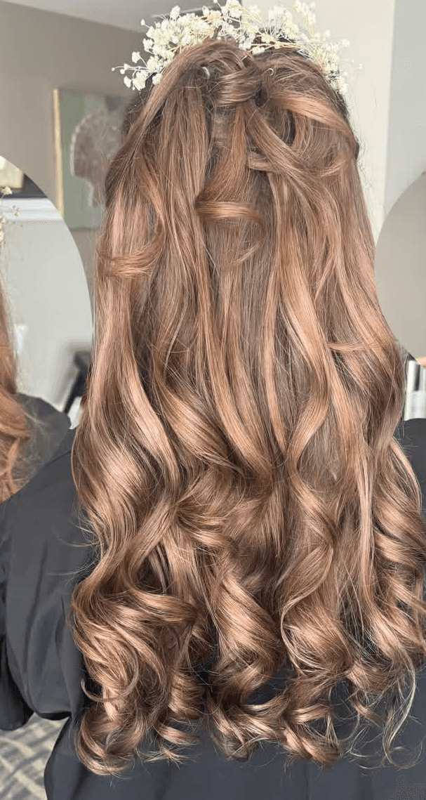 The image shows a rear view of a finished bridal hairstyle on a client, photographed indoors with soft, natural lighting. The hair is long and styled in loose, defined curls with a half-up structure, featuring subtle twists and interwoven sections that add dimension and structure at the crown. A delicate white floral hairpiece is placed at the top, integrated into the style without obscuring the hair’s texture or movement. The composition focuses tightly on the hairstyle’s shape, curl pattern, and detailing, making it suitable for a bridal portfolio or wedding services gallery on a hairstylist’s website.