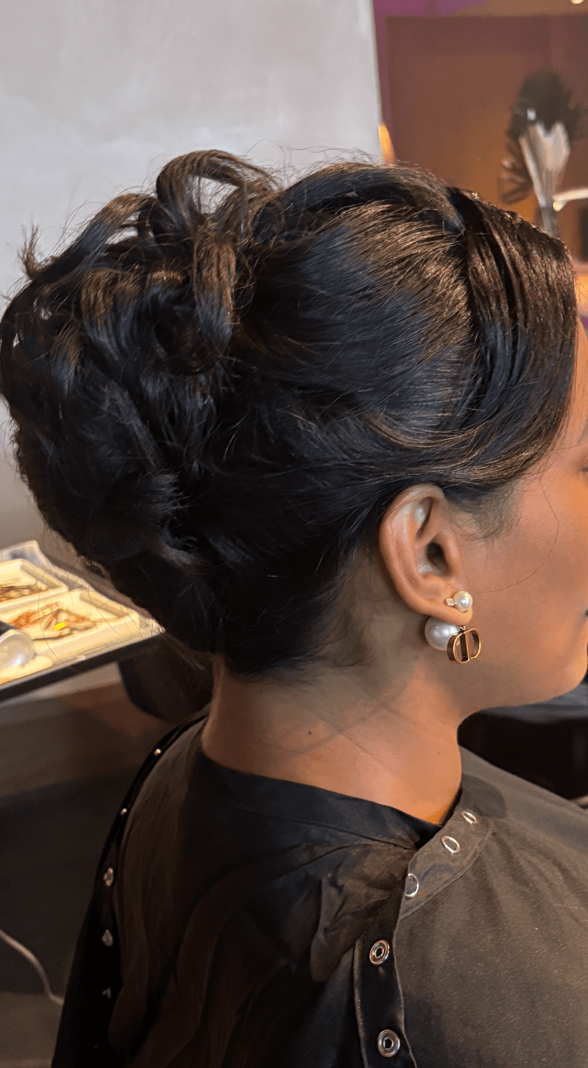 The image shows a side-profile view of a completed formal updo hairstyle on a client seated in a salon chair. The hair is dark and styled into a structured, low bun with woven sections and soft volume, creating a smooth, polished silhouette with visible texture and depth. The client is wearing stud and drop earrings, and a black styling cape, with the hairstyle clearly framed from ear to nape. The background is softly blurred with salon tools and warm indoor lighting, making the image suitable for an updo portfolio, special-event styling, or bridal/formal services section on a hairstylist’s website.