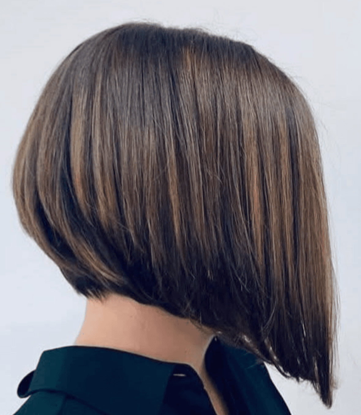 The image shows a side and rear angled view of a precision-cut asymmetrical bob hairstyle against a plain, light background. The haircut features a sharply angled perimeter, shorter at the nape and gradually lengthening toward the front, creating a clean, geometric silhouette. The hair is smooth and straight with subtle, natural-looking highlights blended through a medium brown base, emphasizing depth and movement along the angle. The minimal background and tight framing isolate the cut’s structure and line work, making the image suitable for a haircut portfolio, technical style reference, or featured service image on a hairstylist’s website.